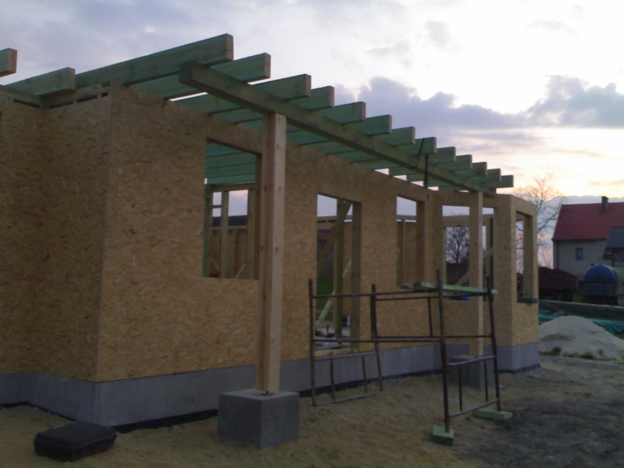 Konstrukcja domu w technologii szkieletowej: ściany z płyt OSB, drewniany szkielet, fundament z bloczków betonowych. Widoczne rusztowanie i elementy konstrukcyjne dachu.