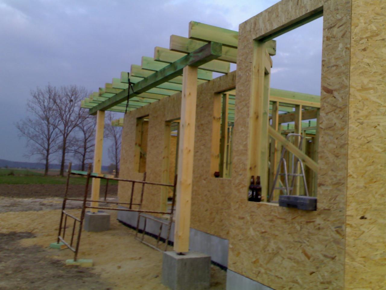 Konstrukcja ścian i dachu z płyty OSB i drewna, widoczne rusztowanie i butelki na parapecie. Budowa w toku.