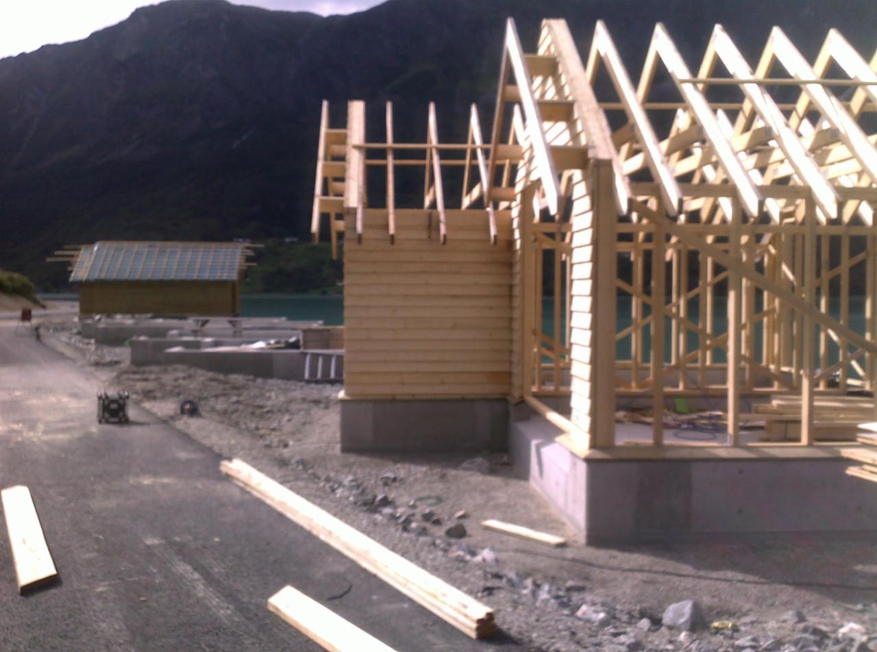 Wznoszenie konstrukcji drewnianej domu na fundamencie, w tle góry i jezioro. Widoczne elementy szkieletu i deski elewacyjne. Etap budowy.
