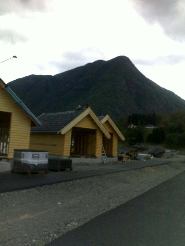 Wznoszenie domów w konstrukcji szkieletowej na tle górskiego krajobrazu. Częściowo wykończone budynki z widoczną konstrukcją dachu i żółtą elewacją. Na dachu pracują dwie osoby.