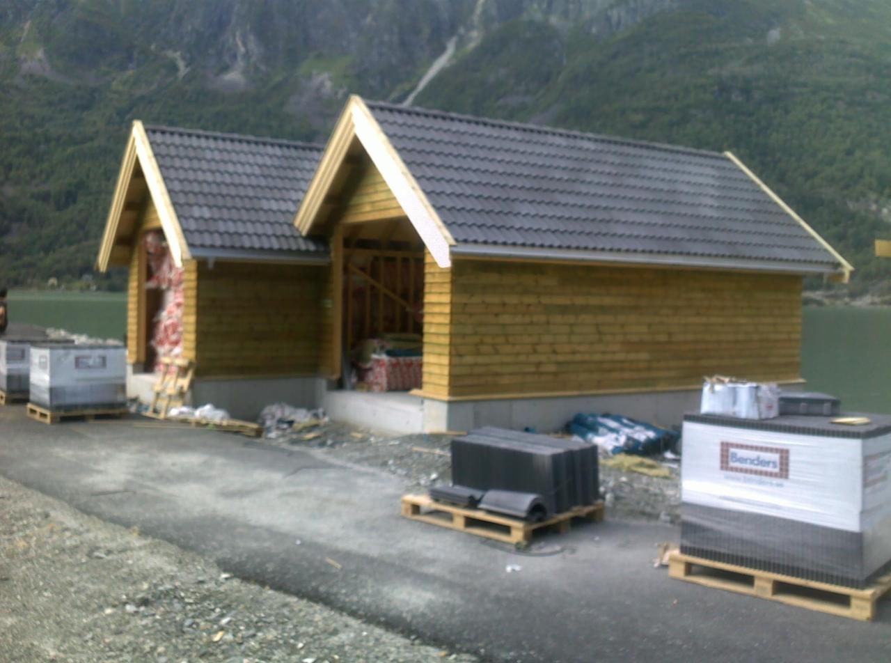 Budowa dwóch małych domów szkieletowych z drewnianą elewacją i szarą dachówką. Materiały budowlane na paletach przed budynkami. W tle góry i jezioro.