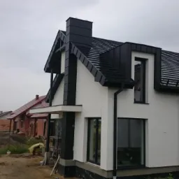 Nowoczesny dom jednorodzinny z czarnym dachem z dachówki ceramicznej i obróbkami blacharskimi. Widoczne rynny i komin.