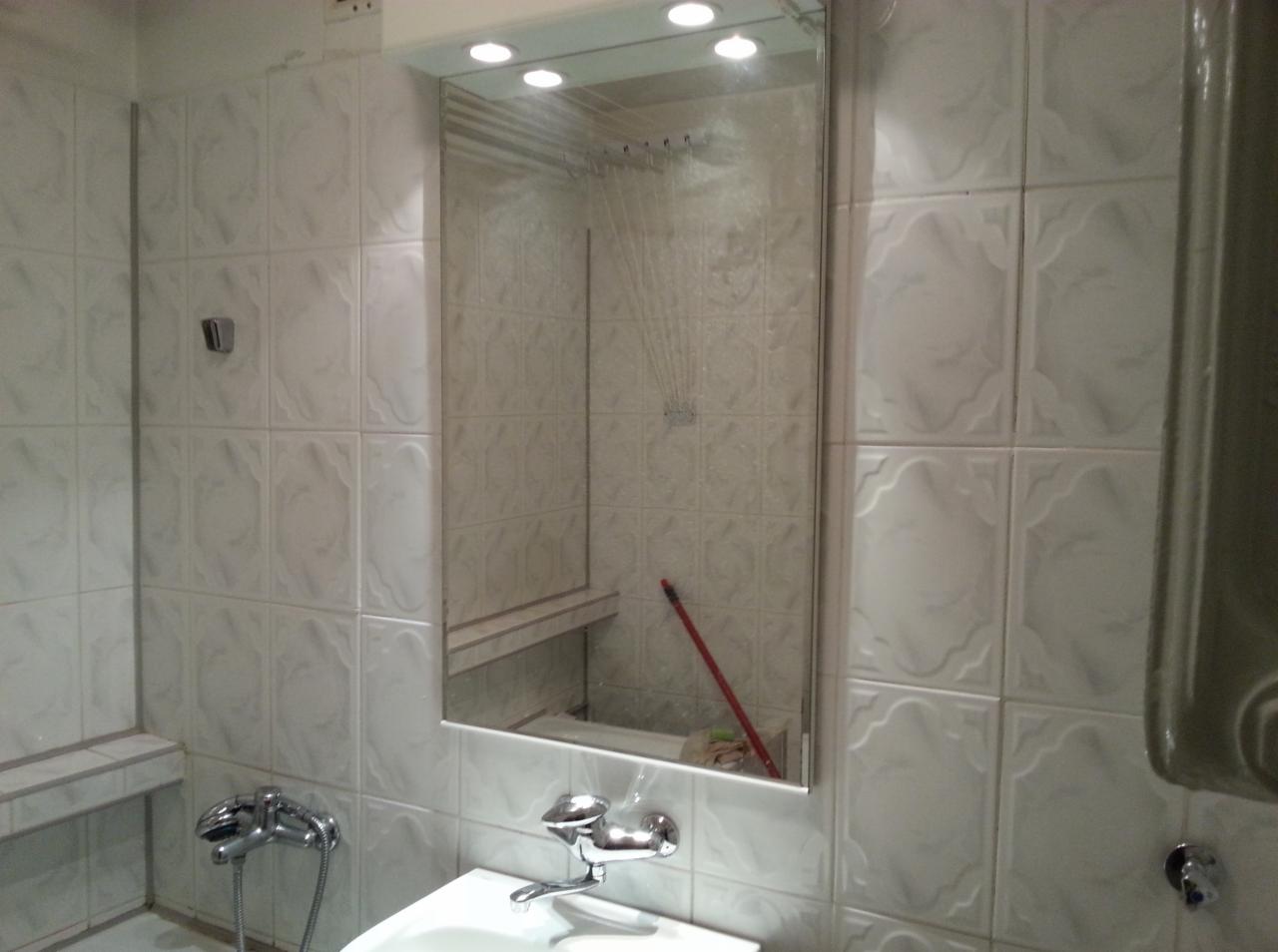 Wykończona łazienka z białymi płytkami i lustrem, w którym odbija się prysznic z suszarką na ubrania. Widoczna umywalka i bateria.