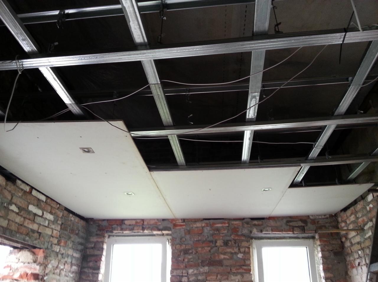 Montaż sufitu podwieszanego z płyt gipsowo-kartonowych na metalowej konstrukcji w pomieszczeniu z ceglanymi ścianami i oknami. Widoczne instalacje.