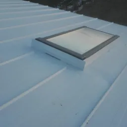 Biały dach z blachy trapezowej z zamontowanym oknem dachowym z szarą ramą, widoczne łączenia blachy.