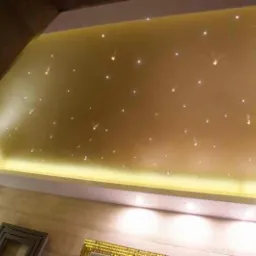 Podwieszany sufit z oświetleniem LED imitującym gwiazdy, w ciepłej tonacji, widok z dołu, fragment ściany w tle.