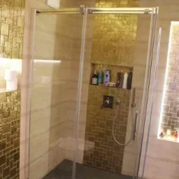 Nowoczesna kabina prysznicowa z transparentnego szkła, złote mozaiki na ścianach, wbudowana półka z kosmetykami, deszczownica sufitowa, minimalistyczny design.