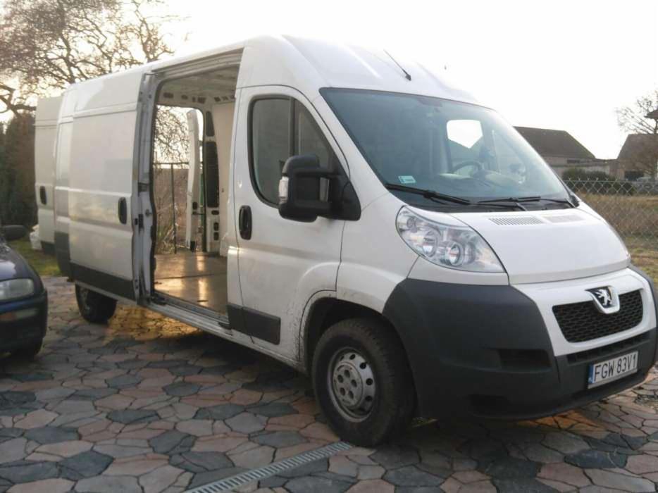 Biały Peugeot Boxer z otwartymi drzwiami bocznymi, ukazujący pustą przestrzeń ładunkową, zaparkowany na brukowanym podjeździe. Widoczny fragment innego pojazdu.