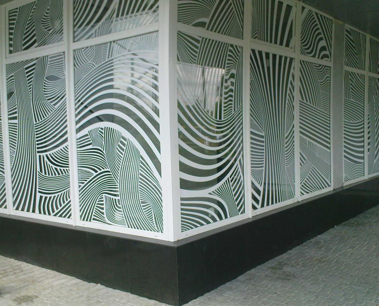 Oklejone witryny sklepowe z abstrakcyjnym wzorem białych linii na szkle. Widok z zewnątrz, kątowe ujęcie. Czarny cokół budynku i szara kostka brukowa.