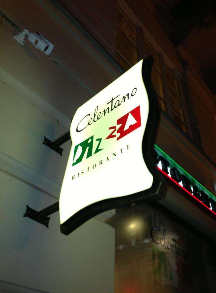 Podświetlany szyld restauracji Celentano Pizza z zielono-czerwonym akcentem, zamontowany na ścianie budynku z widoczną kamerą monitoringu powyżej.