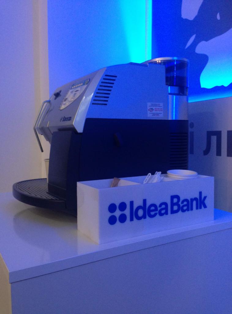 Ekspres do kawy Saeco na białym blacie, obok pojemnik z logo Idea Bank, zawierający mieszadła i kubeczki, całość oświetlona niebieskim światłem.