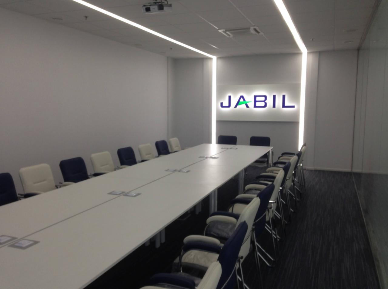 Sala konferencyjna z długim stołem i krzesłami, podświetlane logo Jabil na ścianie, nowoczesne oświetlenie sufitowe, ciemna wykładzina podłogowa.