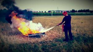 Mężczyzna w czerwonej czapce gasi pożar w metalowej skrzyni na łące, używając czerwonej gaśnicy. Czarny dym unosi się nad płomieniami.