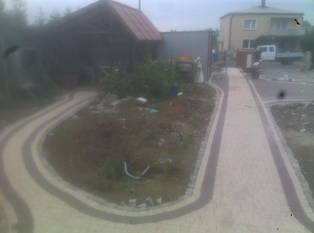 Ścieżka z jasnej kostki brukowej z ciemnoszarym obramowaniem, prowadząca przez plac budowy z betoniarką i stertą ziemi. W tle budynek mieszkalny i zaparkowany samochód dostawczy.