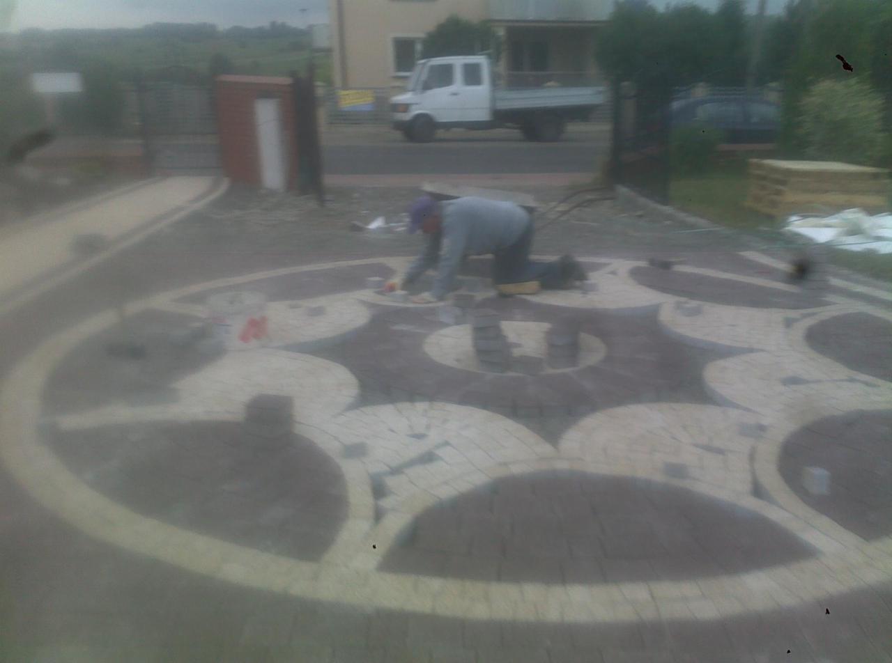 Pracownik w trakcie układania wzoru z kostki brukowej na podjeździe. Widoczne elementy dekoracyjne i częściowo ułożona powierzchnia. W tle budynek i samochód dostawczy.