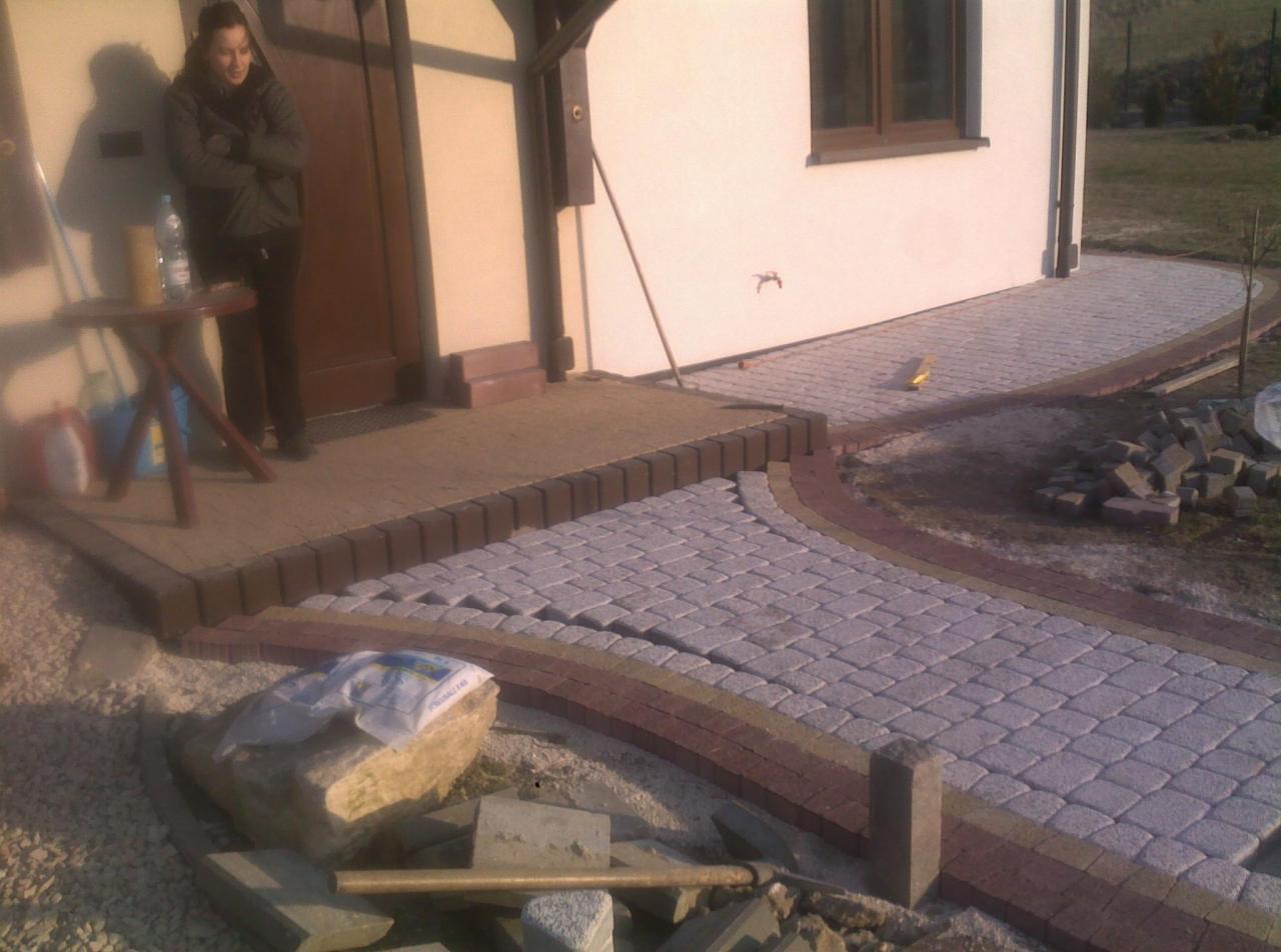 Kaskadowe ułożenie kostki brukowej w odcieniach szarości i brązu, prowadzące od drzwi domu do ogrodu. Widoczna kobieta przy wejściu i narzędzia brukarskie.