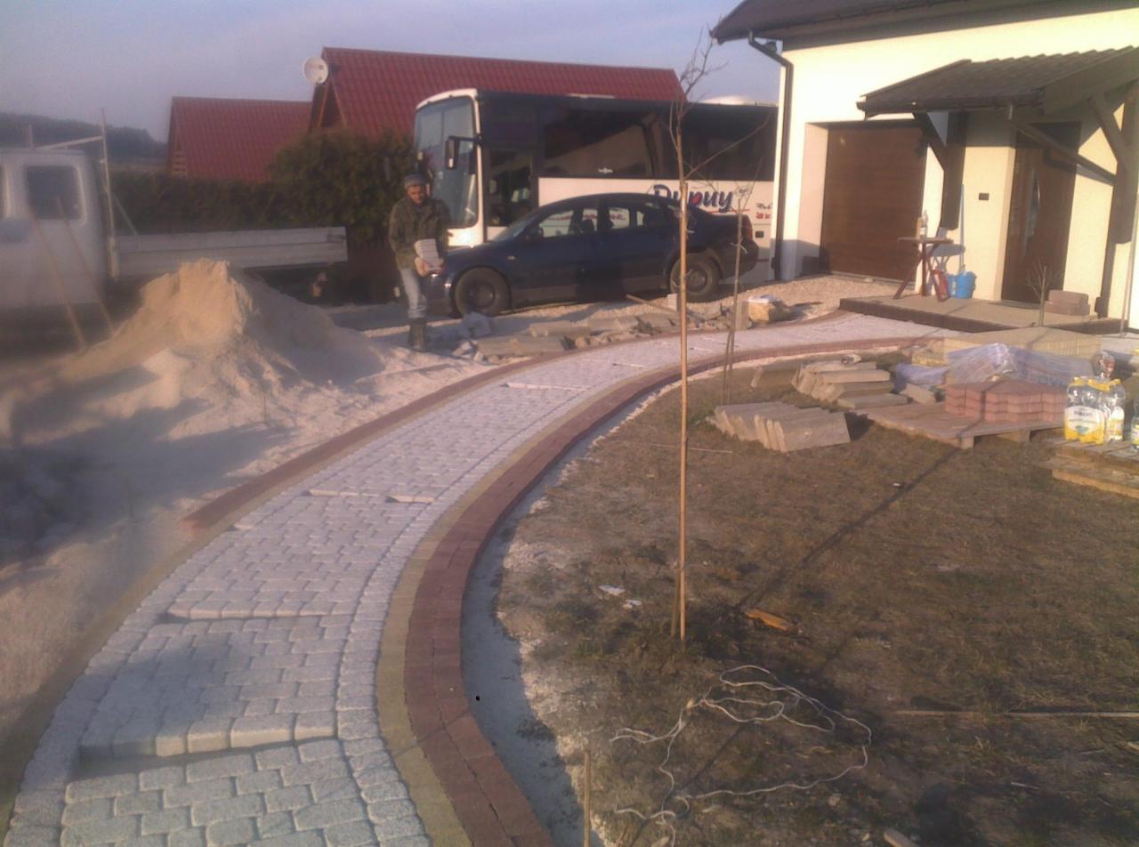 Zakrzywiony chodnik z kostki brukowej w Działoszycach, obrzeża z czerwonej kostki. Na posesji zaparkowany autobus i samochód osobowy, widoczny pracownik z kostką.