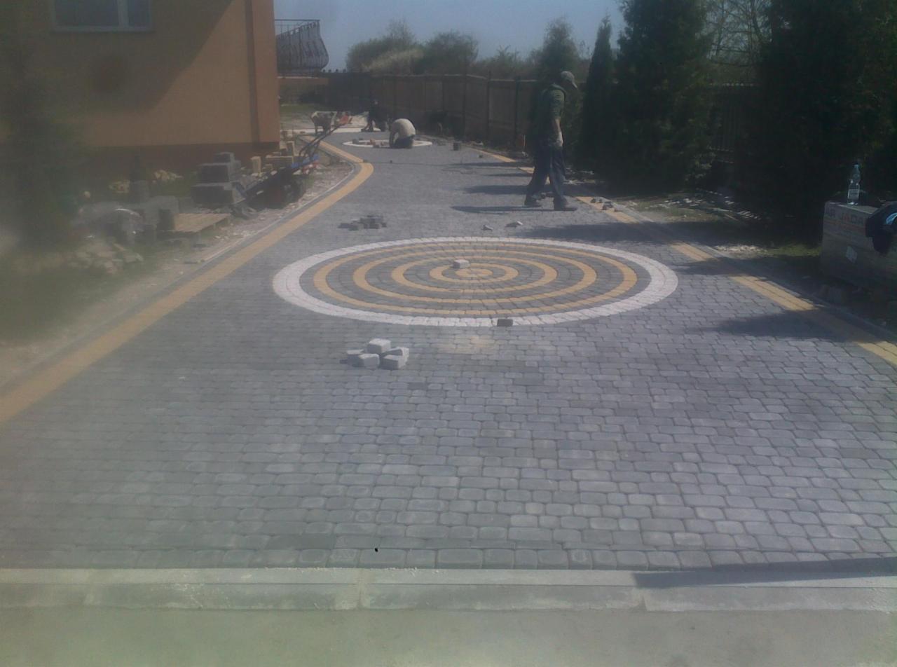 Szara kostka brukowa ułożona w wzór z okręgami w kolorach białym i żółtym. W tle pracownicy i budynek. Częściowo ukończony projekt.