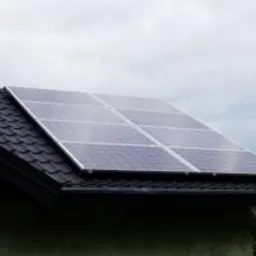 Panele słoneczne zamontowane na dachu domu pokrytym ciemną dachówką, widoczne z boku pod kątem, z kominem w tle i pochmurnym niebem.