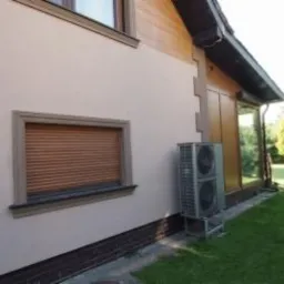 Zewnętrzna jednostka dwuwentylatorowa klimatyzacji/ogrzewania zamontowana na niskim postumencie przy ścianie domu z brązowymi roletami i drewnianą elewacją, widok z boku na trawnik.