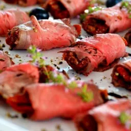 Roladki z rostbefu wypełnione suszonymi pomidorami i bazylią, udekorowane jagodami i ziołami na białym talerzu.