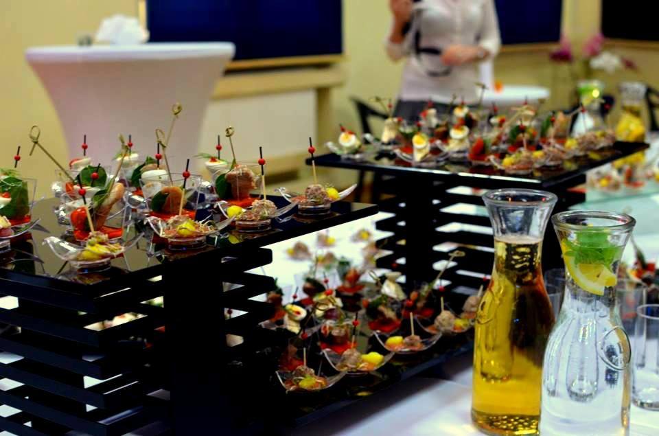 Elegancki catering z przekąskami na czarnych, nowoczesnych stojakach, uzupełniony dzbankami z napojami, tworzący wyrafinowaną aranżację stołu.