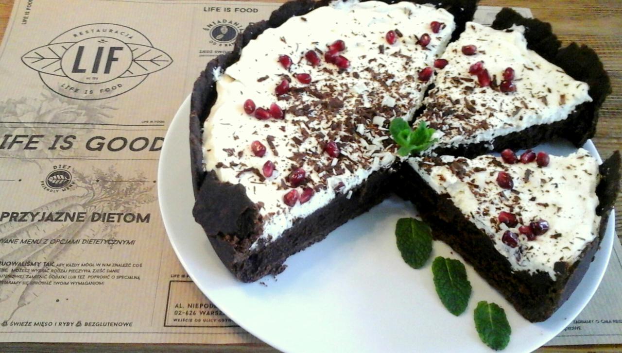 Tort czekoladowy z kremem, posypany startą czekoladą i granatem, udekorowany listkami mięty, na białym talerzu, obok menu restauracji LIF.