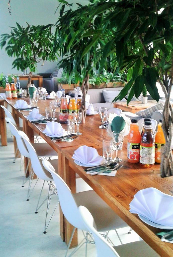 Ustawiony stół z drewnianym blatem, białymi krzesłami, nakryciami, napojami w butelkach i dekoracjami roślinnymi w tle, przygotowany na event.