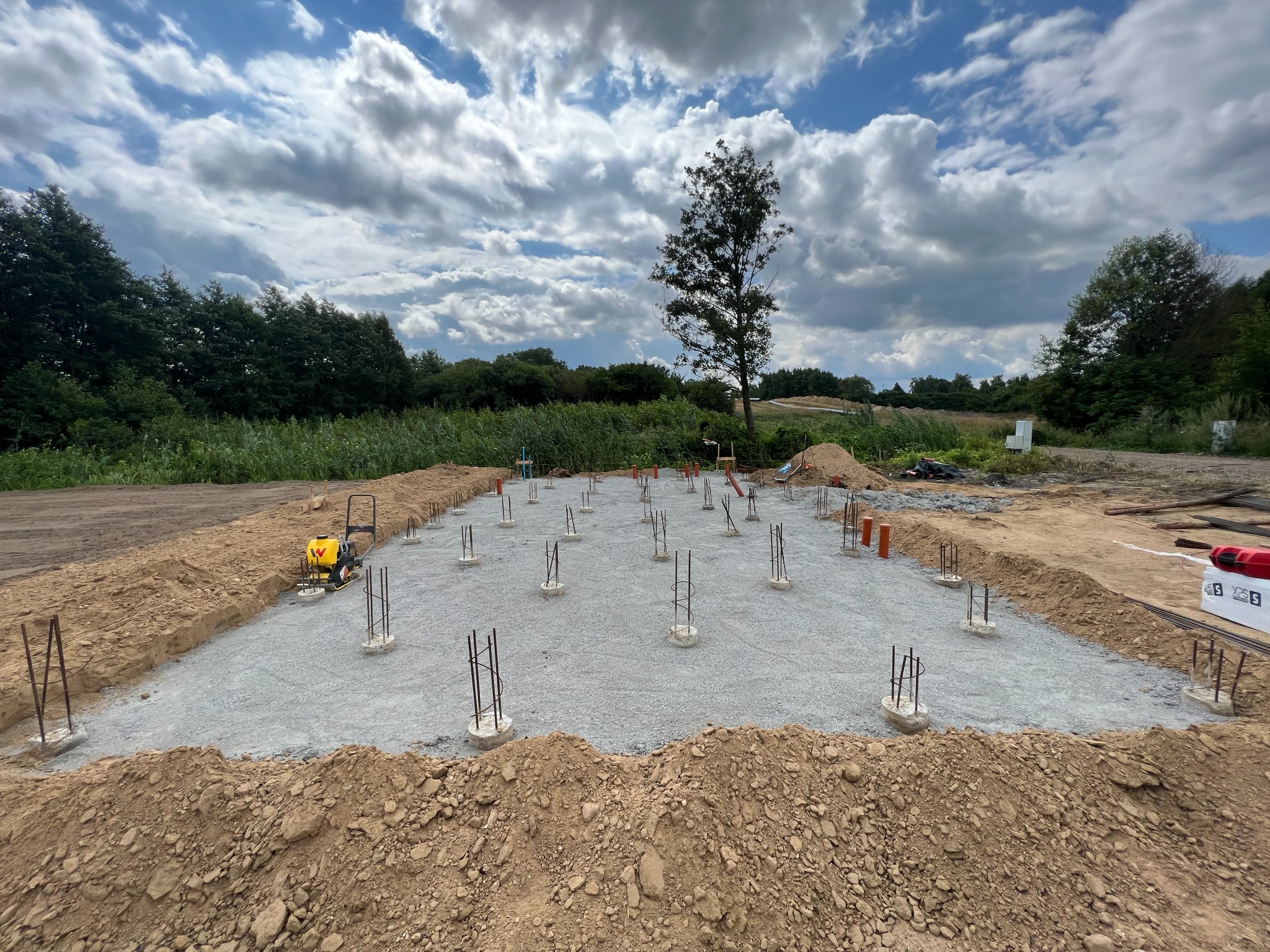Przygotowany teren pod budowę: wyrównany żwir, wystające pręty zbrojeniowe, ubijarka wibracyjna, pomarańczowe rury, widok na zielone drzewa i pochmurne niebo w tle.