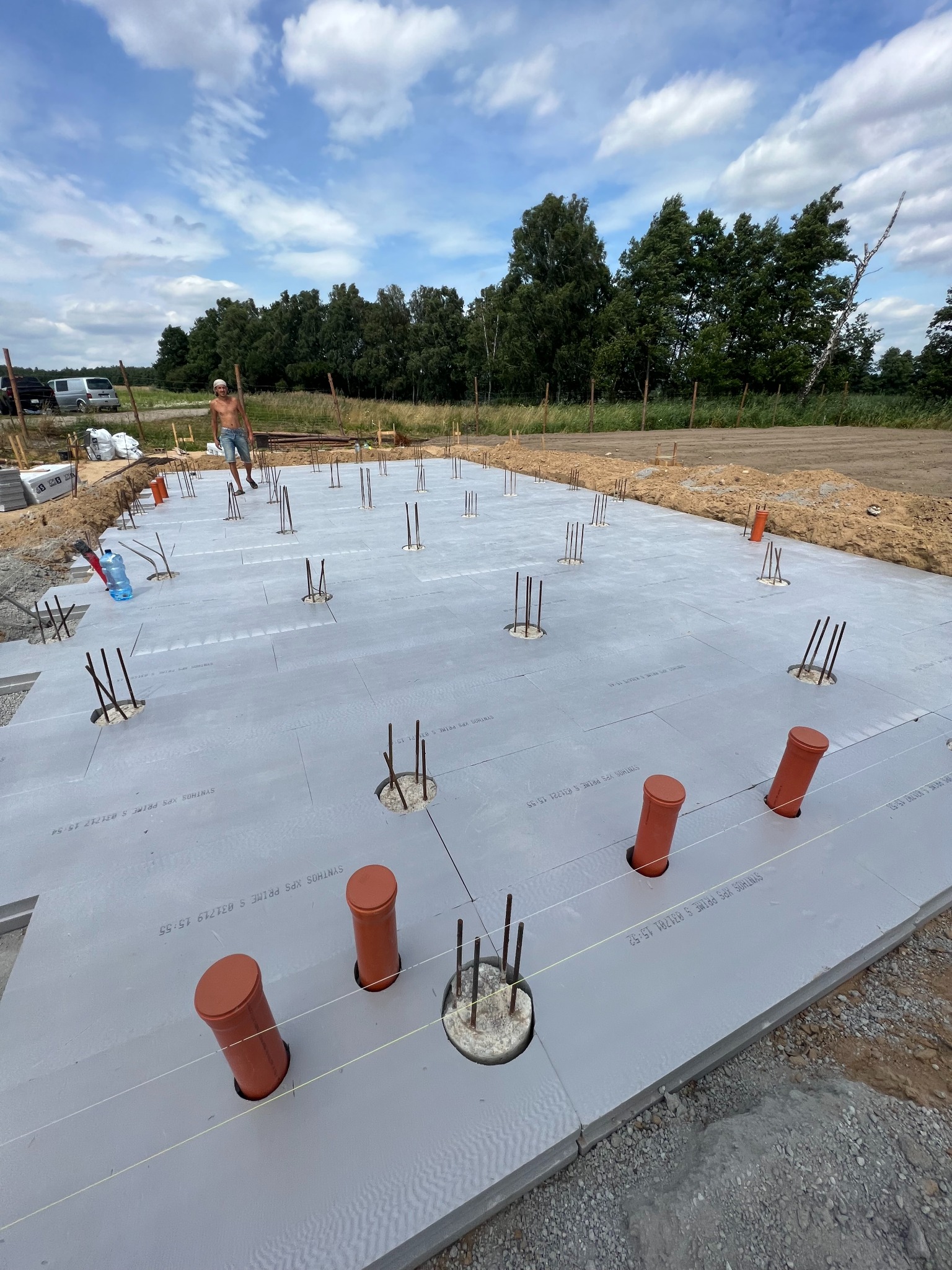 Płyta fundamentowa izolowana styropianem z wystającymi prętami zbrojeniowymi i pomarańczowymi rurami kanalizacyjnymi, widoczny pracownik w tle, teren budowy w słoneczny dzień.