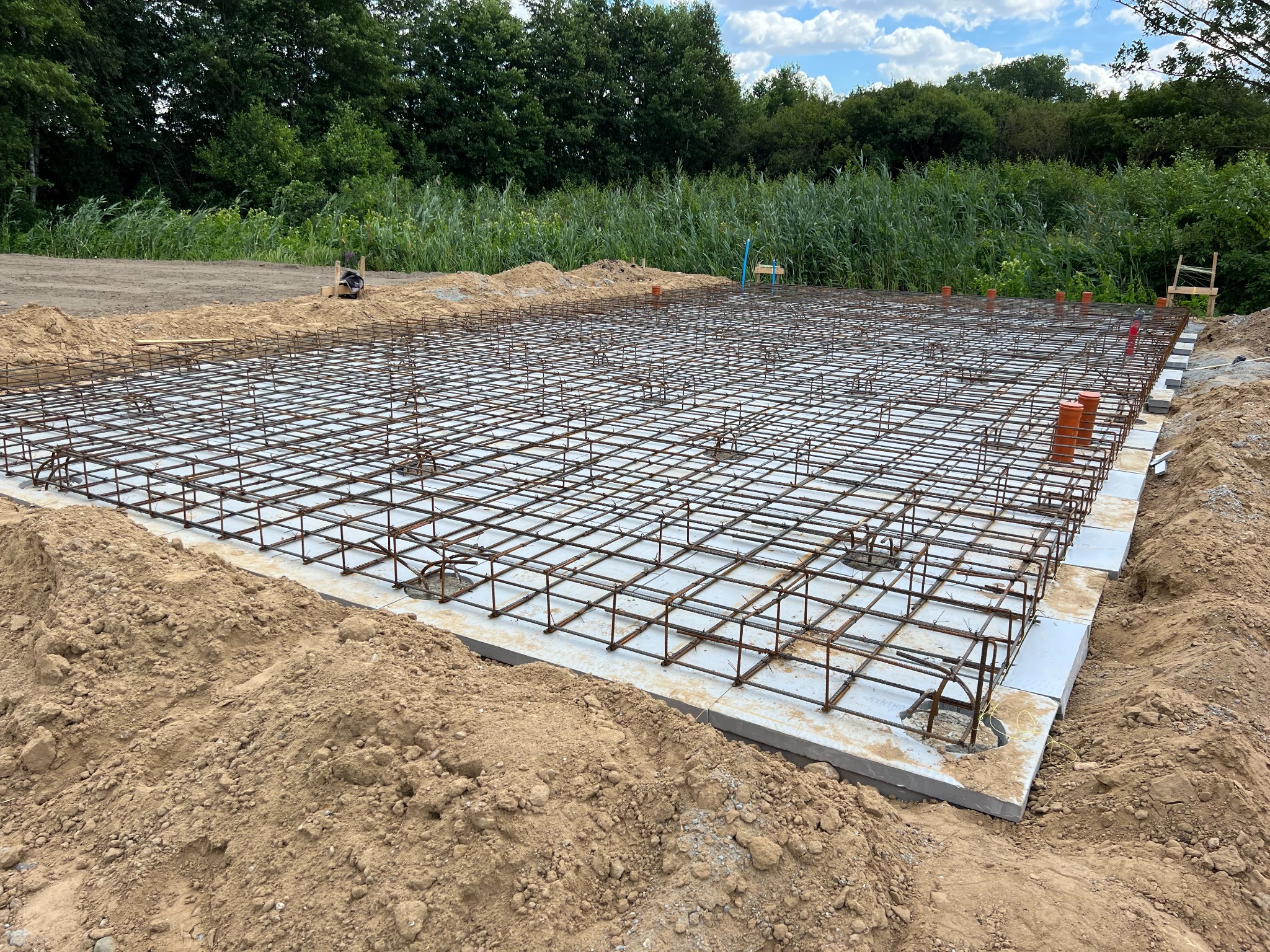 Zbrojenie płyty fundamentowej z ułożonymi bloczkami szalunkowymi, widoczne wystające rury kanalizacyjne i otaczający grunt, w tle zieleń drzew i krzewów.