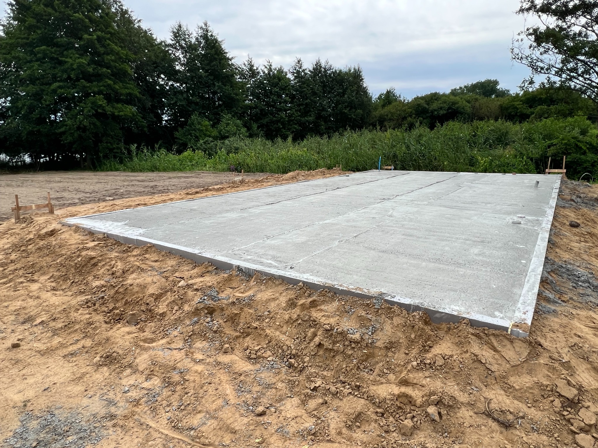 Wylany betonowy fundament na przygotowanym podłożu z widocznymi śladami po deskowaniu, w tle zieleń drzew i krzewów pod pochmurnym niebem.