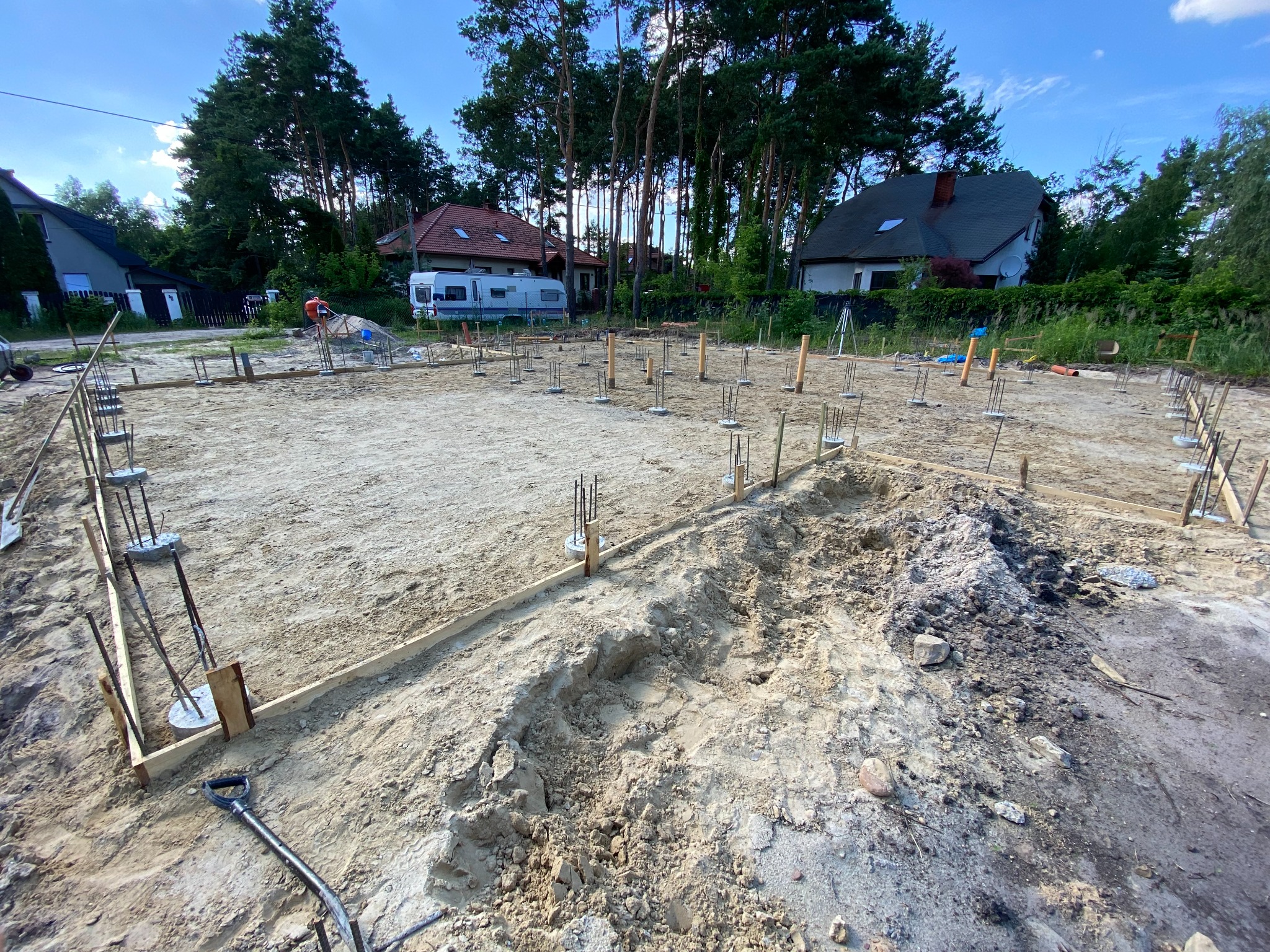 Przygotowany teren pod budowę domu jednorodzinnego z widocznymi szalunkami i wystającymi prętami zbrojeniowymi, w tle domy i drzewa.