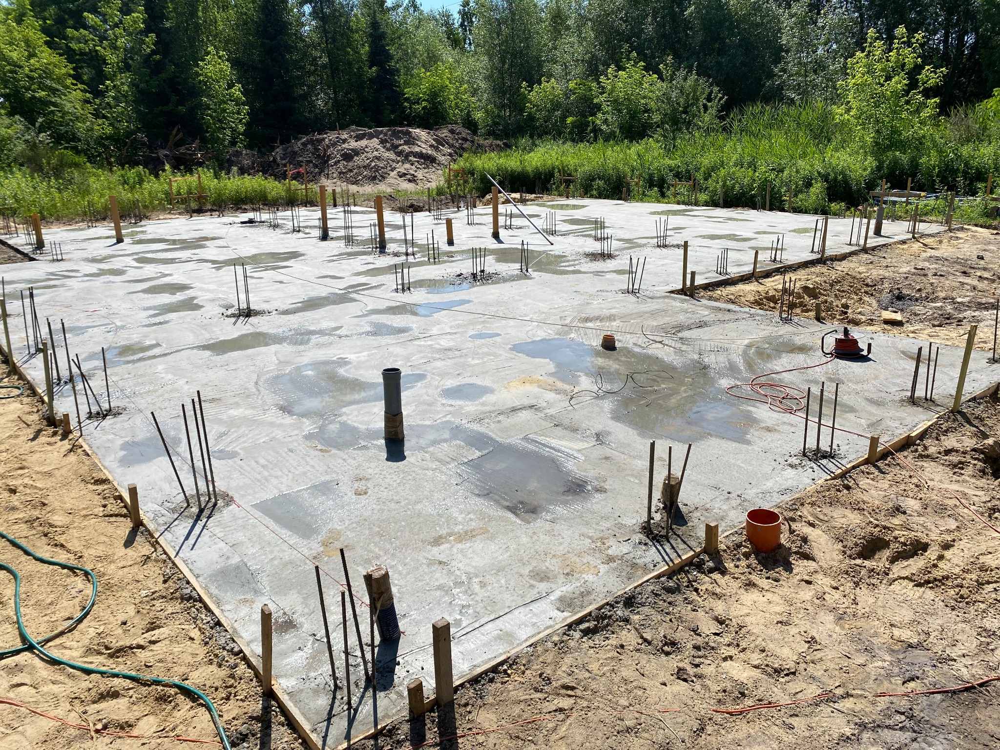 Świeżo wylany betonowy fundament z wystającymi prętami zbrojeniowymi, drewnianym szalunkiem i elementami instalacji, widok z góry na plac budowy w słoneczny dzień.