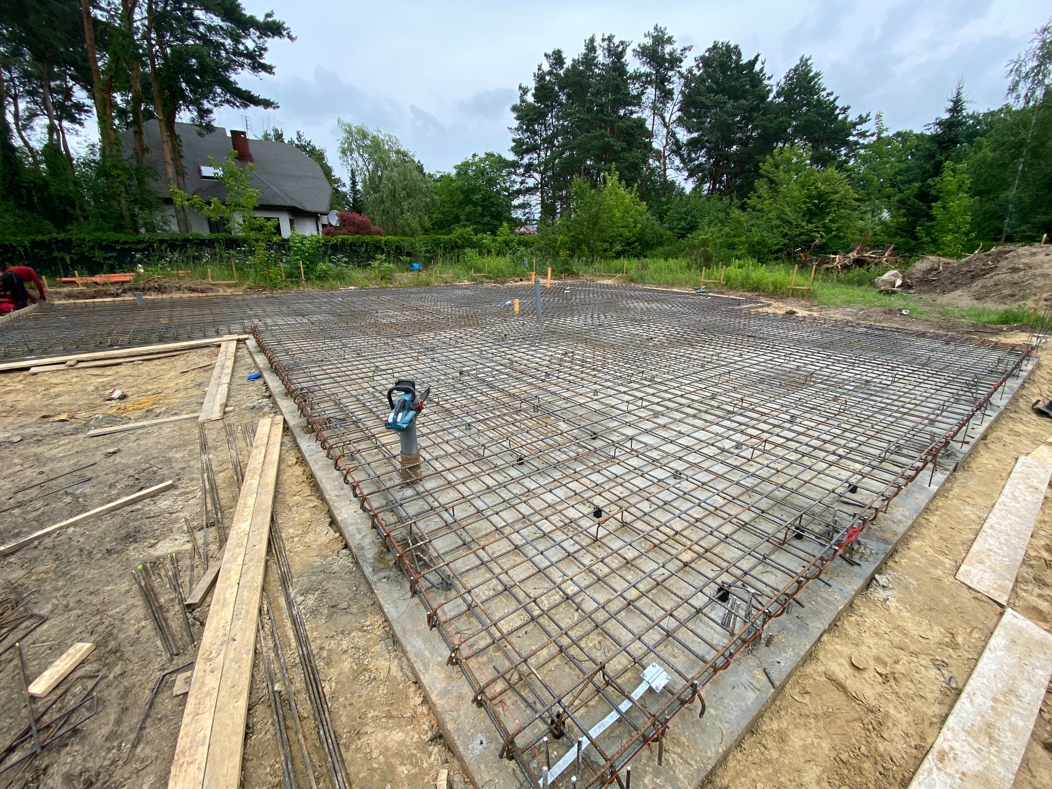 Zbrojenie płyty fundamentowej z wystającymi rurami kanalizacyjnymi na tle działki z domem i drzewami.