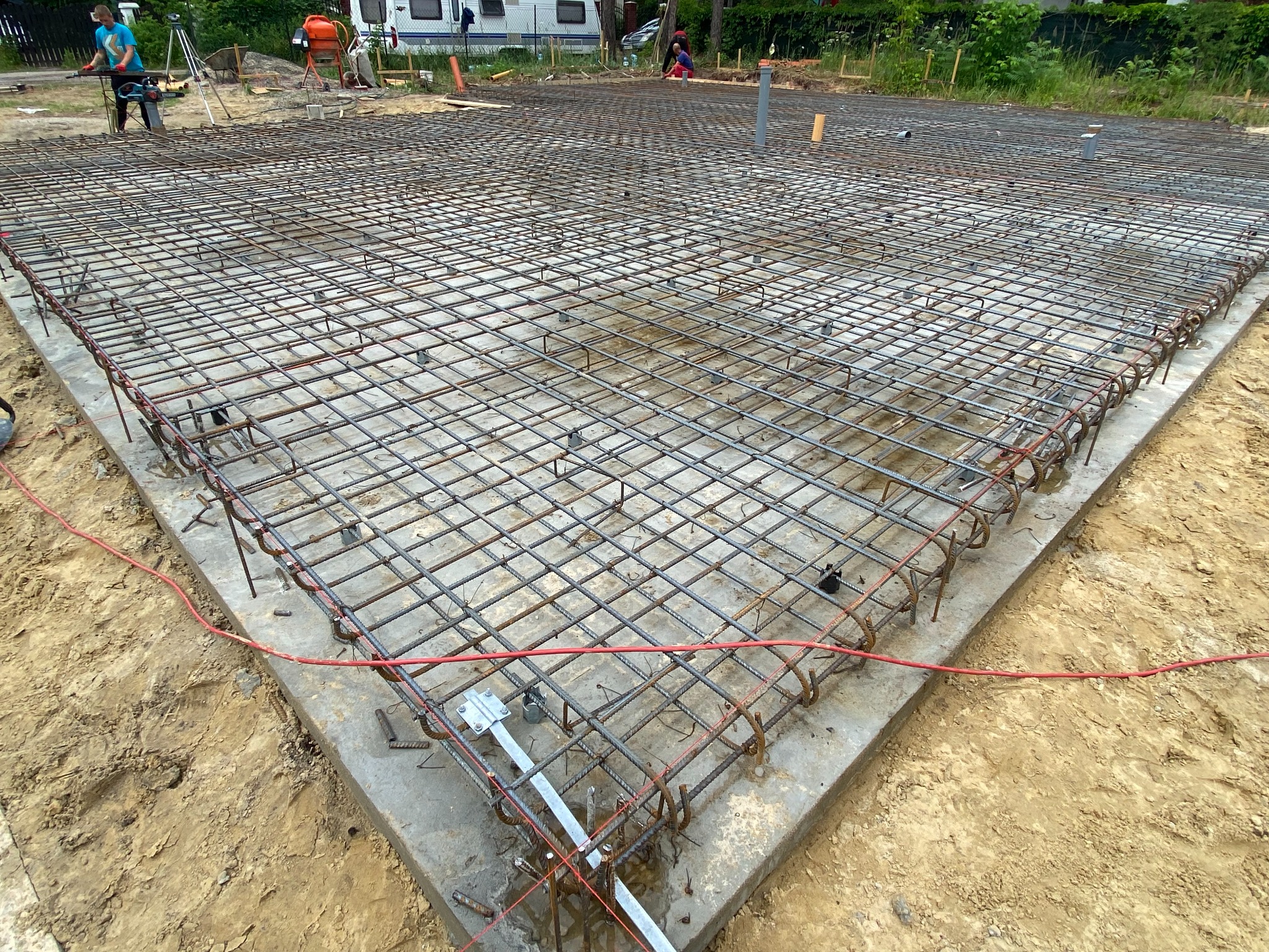 Siatka zbrojeniowa ułożona na betonowej płycie fundamentowej, widoczne wystające pręty, w tle pracownicy budowlani i sprzęt