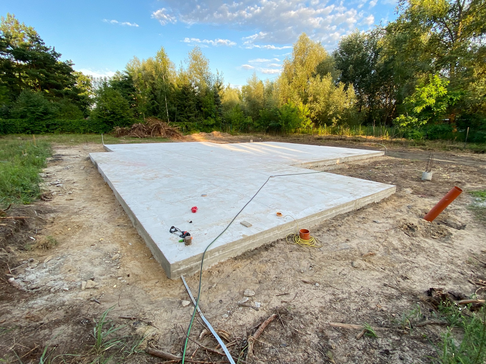 Wylany betonowy fundament płyty z wystającymi rurami instalacyjnymi, widok z poziomu gruntu na tle drzew i błękitnego nieba z chmurami.