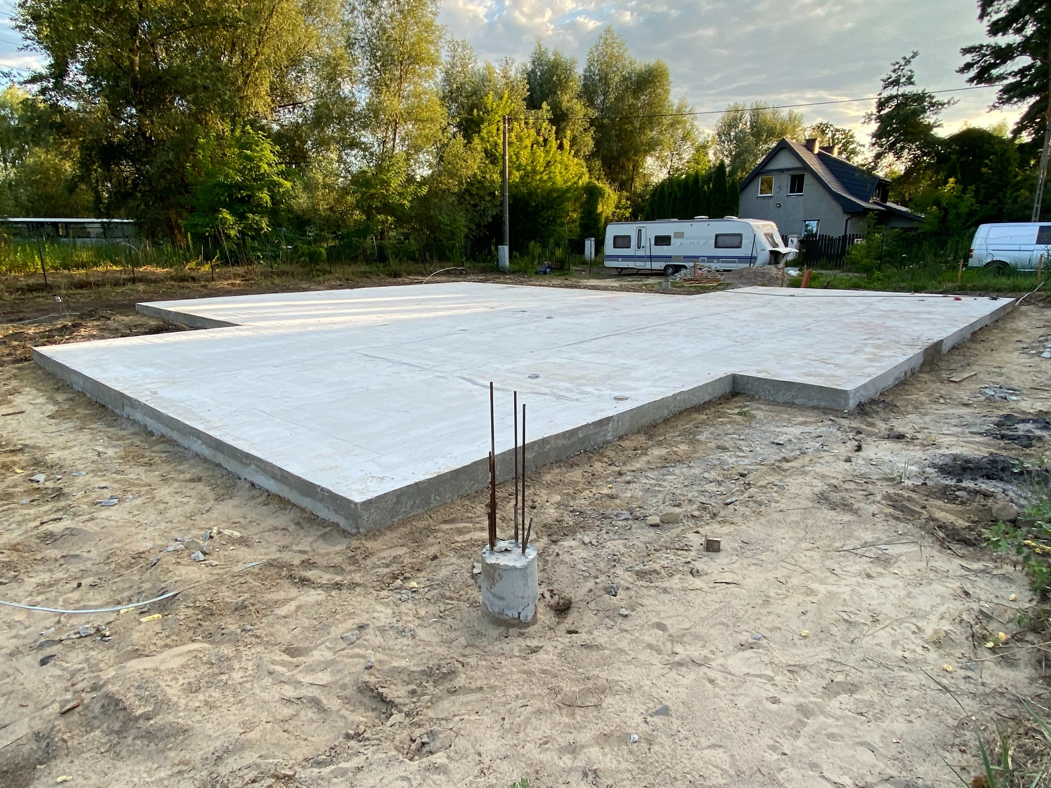 Wylany fundament pod budynek jednorodzinny z wystającymi prętami zbrojeniowymi, na działce widoczny kamper i dom w tle.