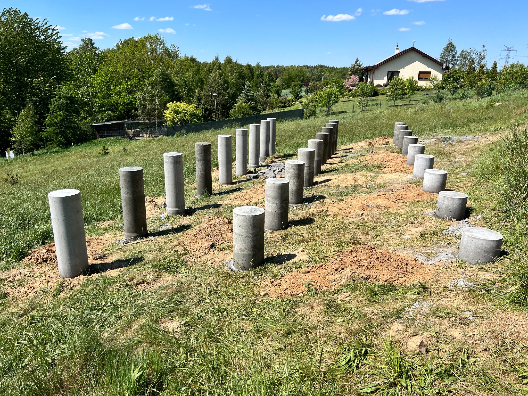 Przygotowane betonowe słupy na gruncie pod budowę domu jednorodzinnego, widok z perspektywy trawnika, dom w tle.