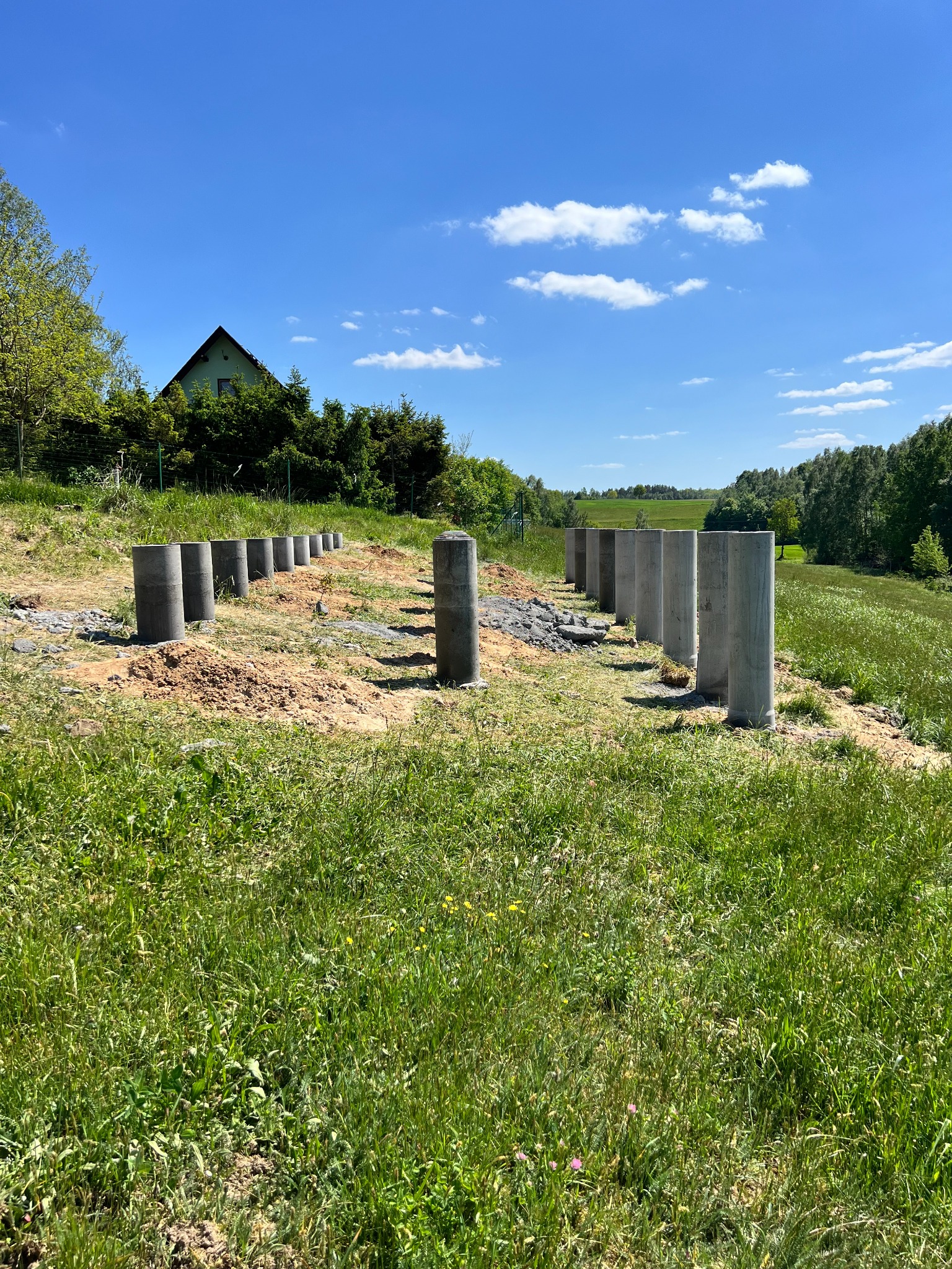 Betonowe słupy przygotowane pod fundamenty na zielonym terenie z widokiem na dom i drzewa w tle, w słoneczny dzień.