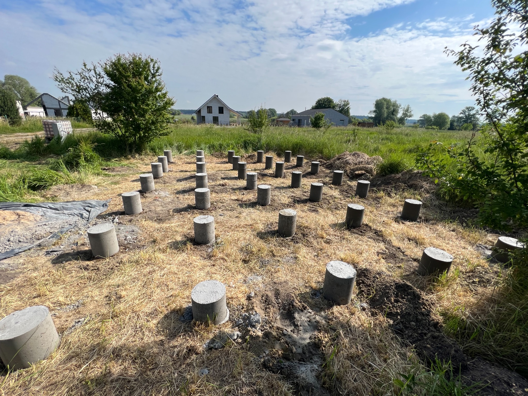 Betonowe słupy fundamentowe na przygotowanym terenie pod budowę domu jednorodzinnego, widoczne w oddali budynki w trakcie budowy.