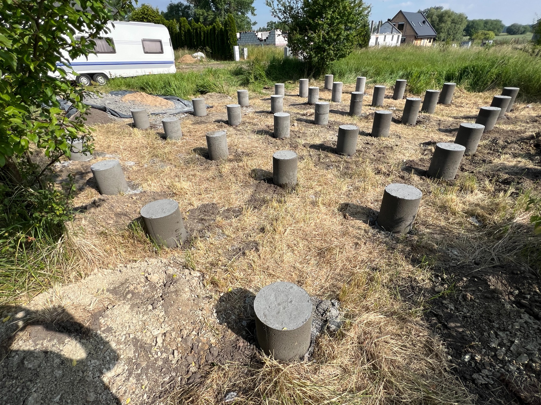 Betonowe słupki fundamentowe na przygotowanym terenie z przyczepą kempingową w tle, widok z poziomu gruntu.