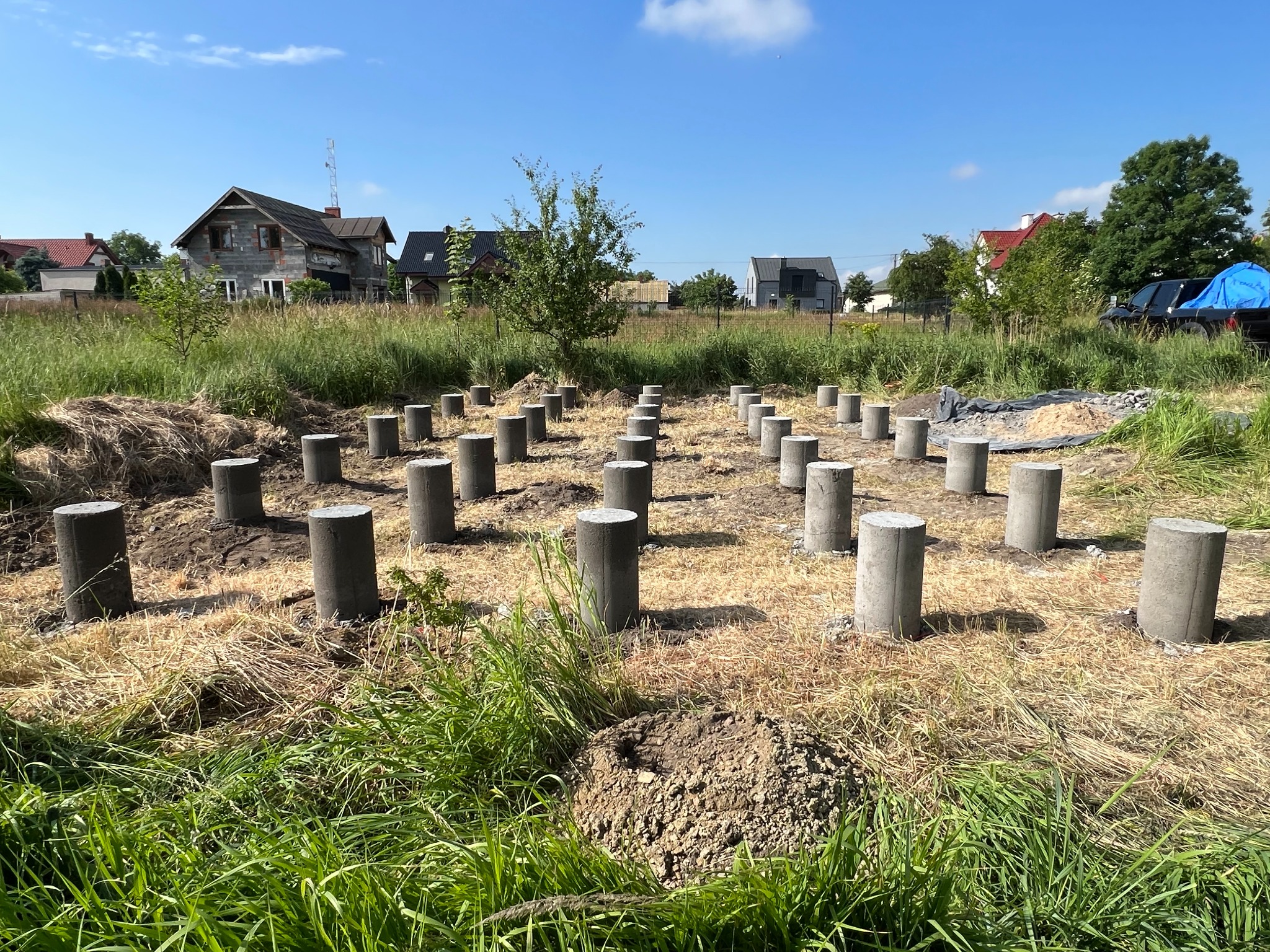Przygotowane betonowe słupy fundamentowe na zalesionym terenie pod budowę domu jednorodzinnego, widoczne domy w tle.