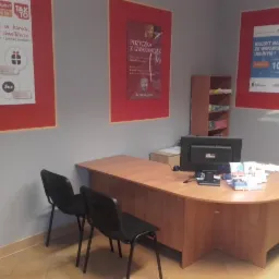 Biuro kredytowe: biurko z komputerem, ulotki i plakaty z ofertami pożyczek na ścianach. Dwa krzesła czekają na klientów. Oświetlenie sufitowe.