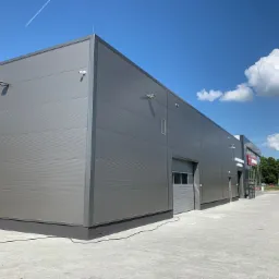 Nowoczesna hala magazynowa z szarej płyty warstwowej, widok zewnętrzny z kostki brukowej, błękitne niebo z chmurami w tle.