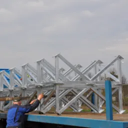 Transport elementów stalowych konstrukcji kratownicowej na niebieskiej platformie ciężarówki, obsługiwane przez zielony wózek widłowy Komatsu, z pracownikiem w niebieskiej kurtce sprawdzającym...