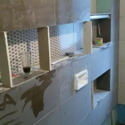 Ściana w trakcie układania płytek z widocznymi wnękami, resztkami zaprawy i szklanką z ciemnym płynem na blacie.