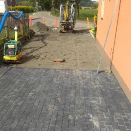 Częściowo ułożona, czarna kostka brukowa, zagęszczarka wibracyjna, mała koparka i przygotowane podłoże z piasku na posesji przy pomarańczowym budynku.