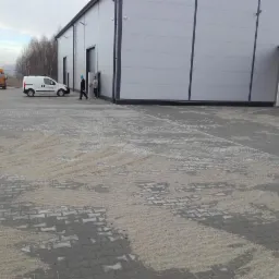Szara kostka brukowa przed budynkiem przemysłowym, częściowo zasypana piaskiem. W tle zaparkowane pojazdy i dwaj pracownicy w pobliżu wejścia.