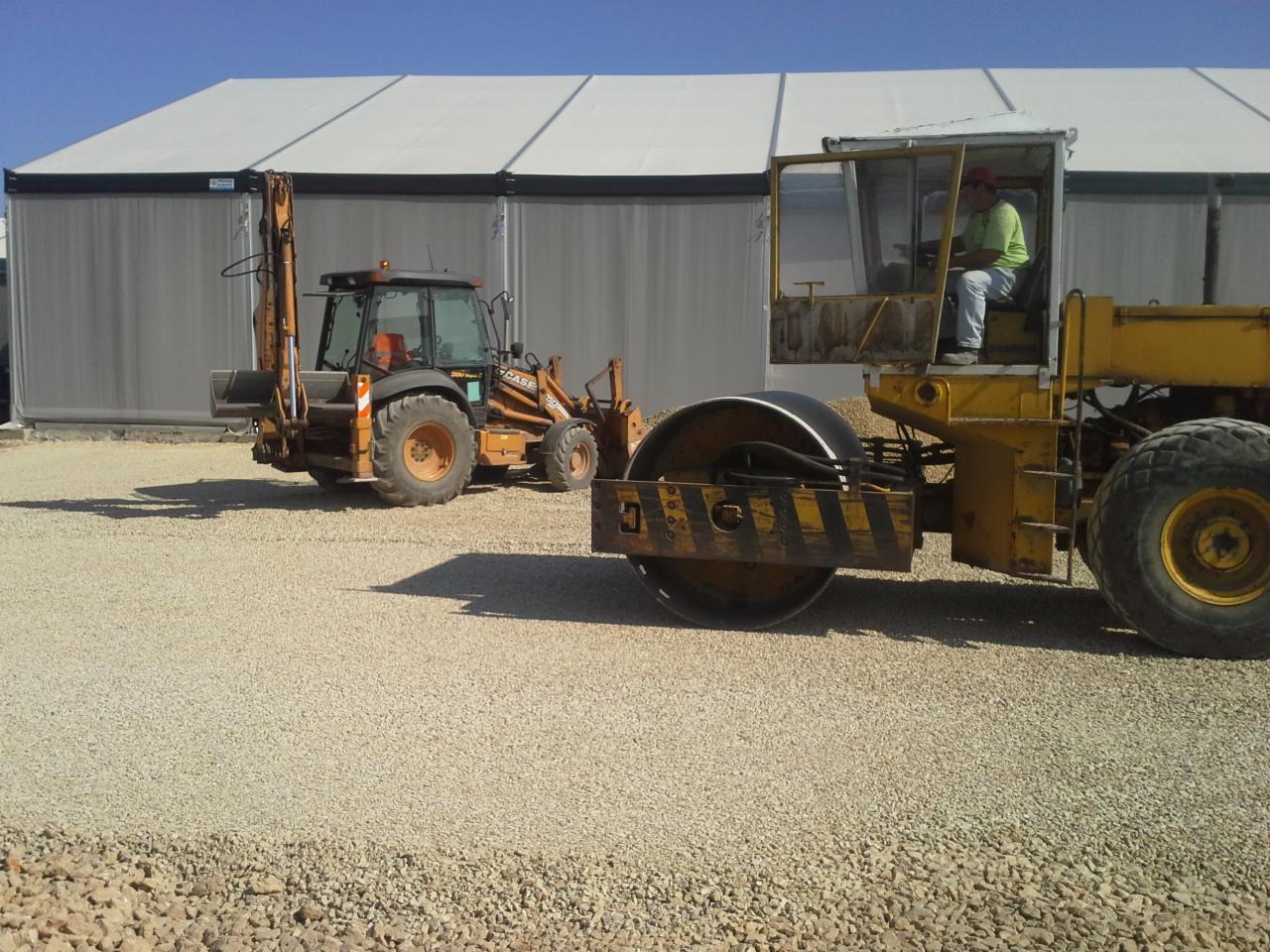 Żółty walec drogowy z operatorem i koparko-ładowarka na żwirowym podłożu. W tle biały, namiotowy budynek.